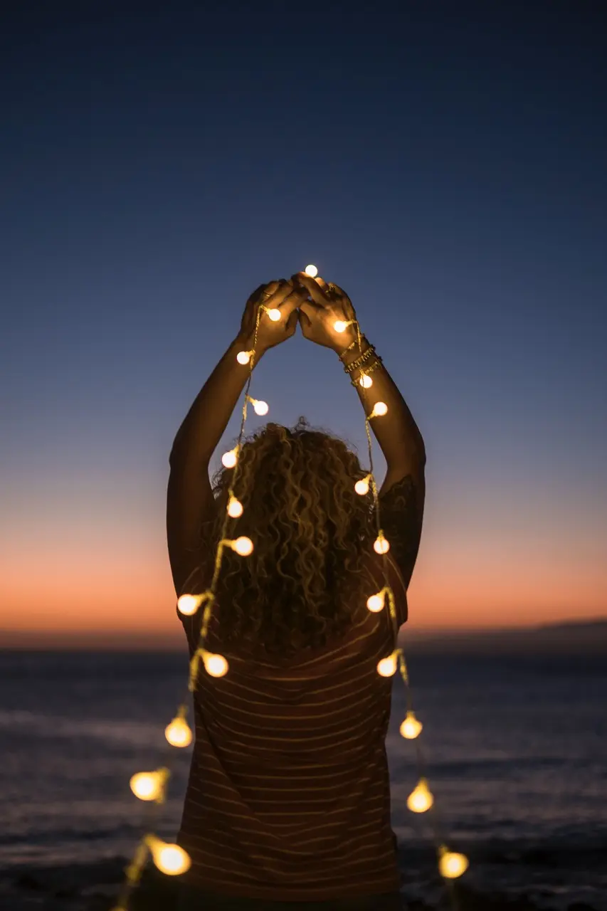 A woman holding string lights at dusk on the beach