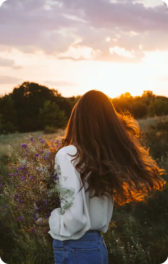 A brunette lady standing in a field of flowers at sunset with a bouquet of flowers in her arms