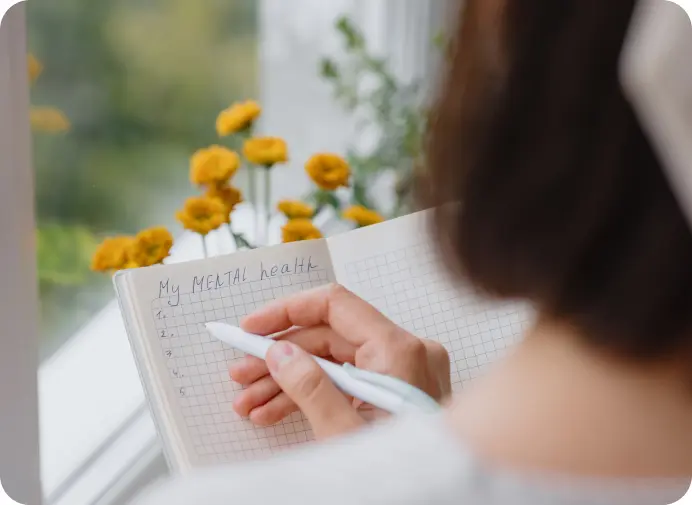 A lady sitting by the window writing in her notebook. Her notebook has the title Mental Health.