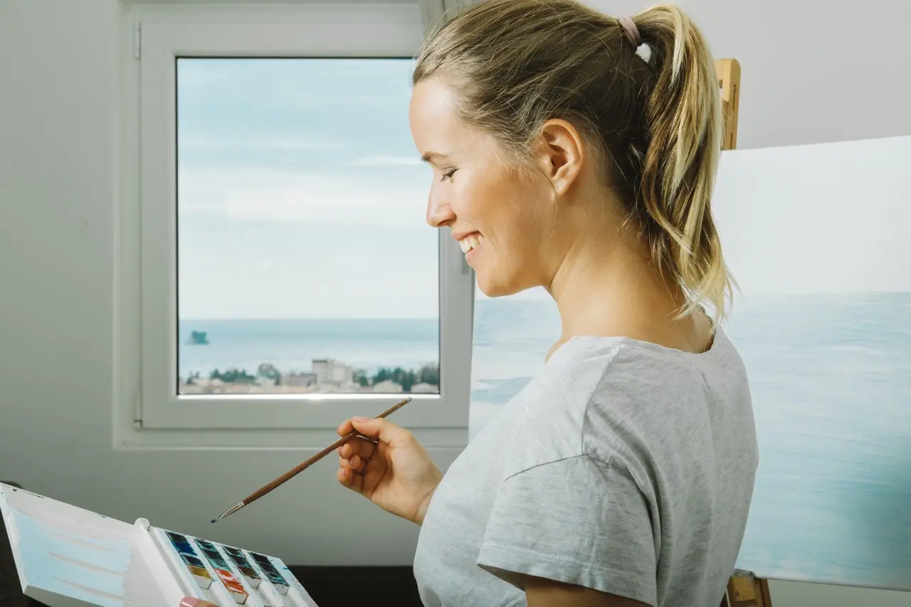 A smiling young woman is painting by the ocean