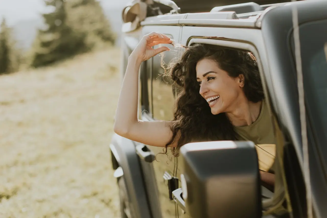 A young woman riding in the passenger front seat of a vehicle, sticking her head and arm out looking happy and free