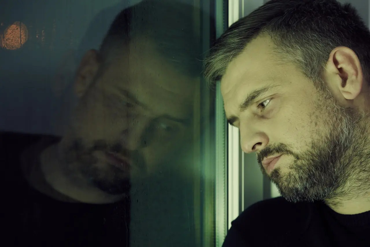 A disheveled man sitting by the window going through an alcohol detox by himself.