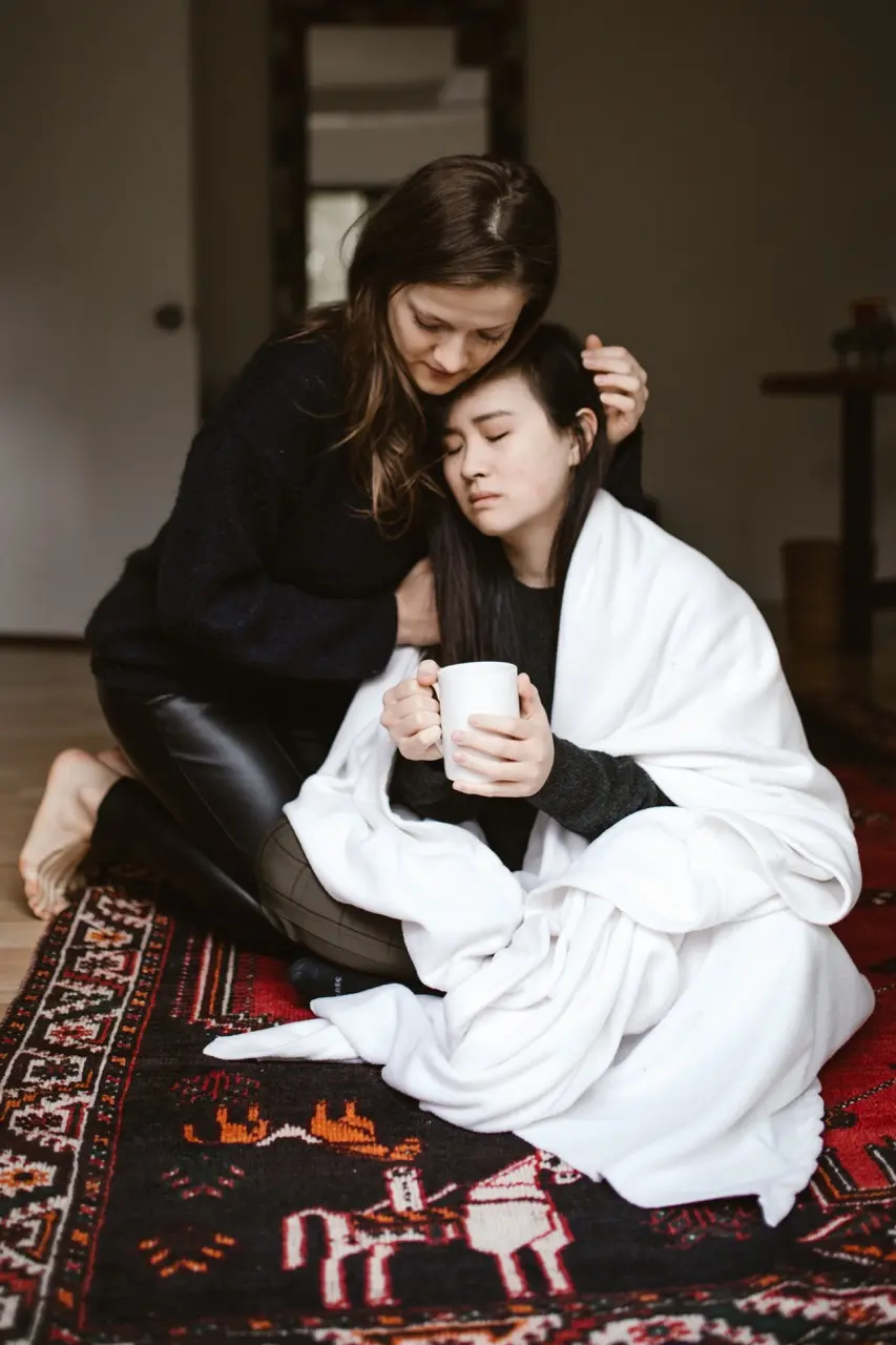 Asian woman suffering from PTSD is wrapped in a white blanket and holding a warm mug, while her friend hugs her in comfort as they both sit on the floor