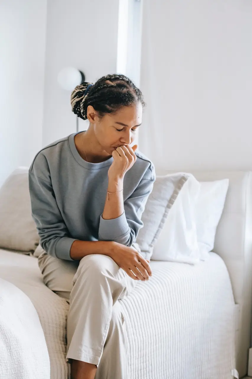 Woman in a blue sweater suffering from anxiety disorder sitting on a white bed with her legs crossed and her chin resting on her fist