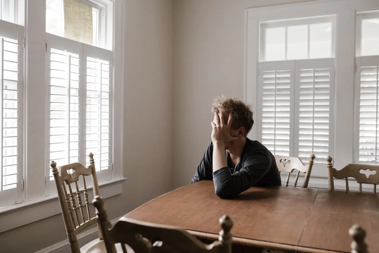 Disheveled man suffering from anxiety disorder sits at a dining table looking out the window with his head resting on his hand.